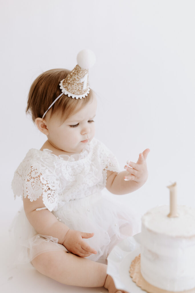 One-year-old baby girl in a lace dress and glittery party hat, sits beside a white smash cake looking at her frosting covered hand during her first birthday milestone session with a Roseville, CA photographer.