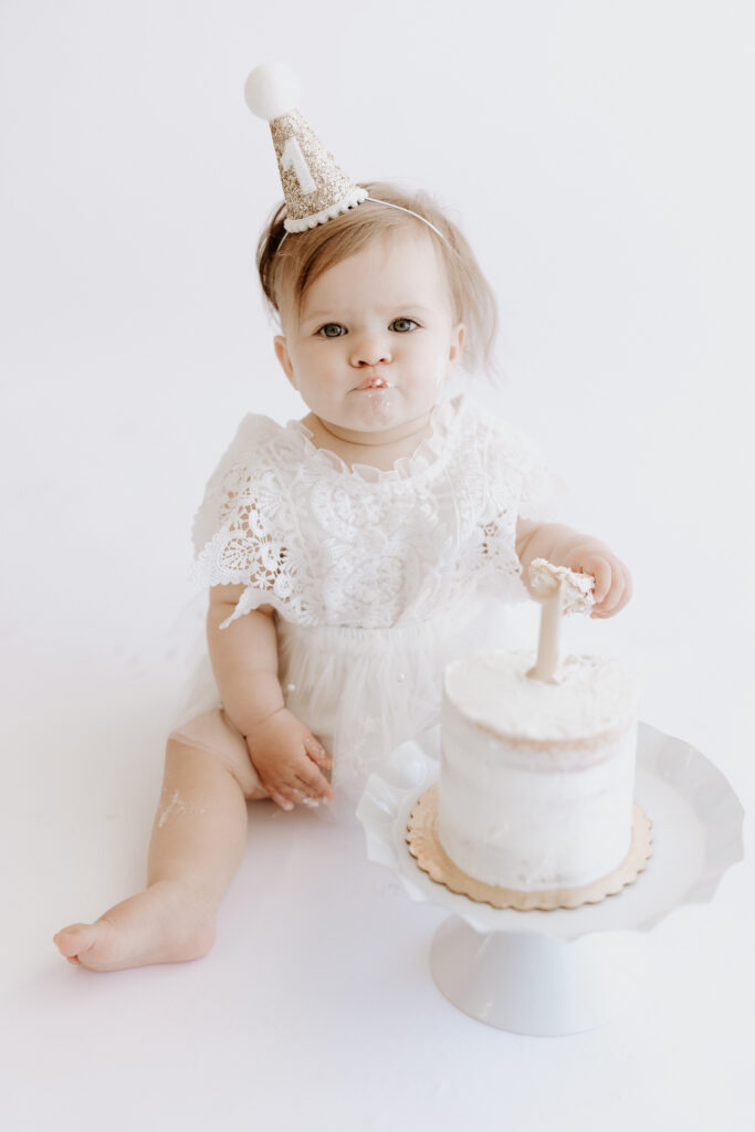 One-year-old baby girl in a lace dress and glittery party hat, sitting beside a white smash cake during her first birthday milestone session with a Roseville, CA photographer.