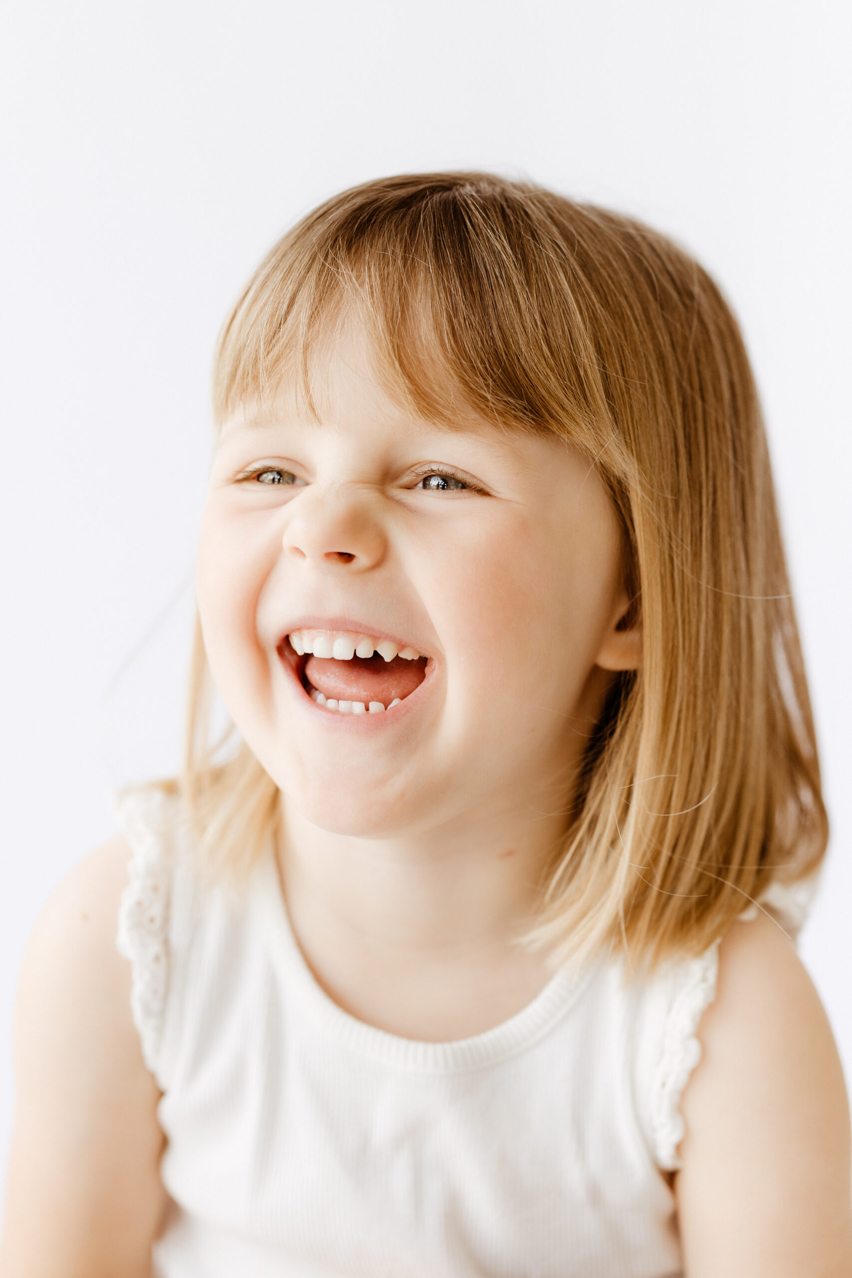 A young girl with straight, light brown hair and wearing a white tank top laughs off camera at her personality portraits session in Roseville, CA.