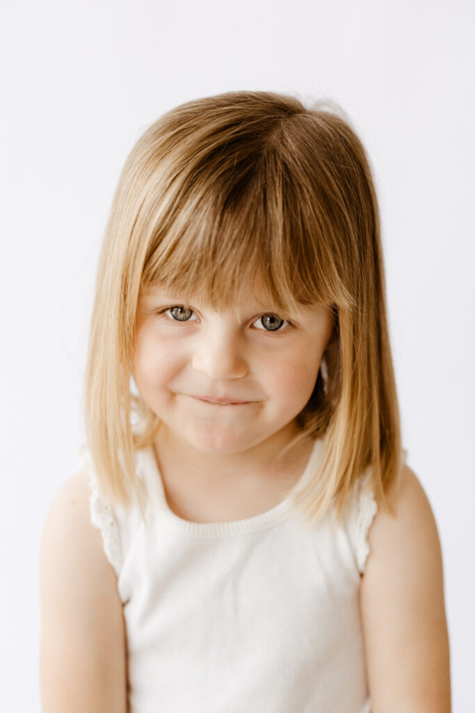Young girl with straight, light brown hair and bangs, wearing a white sleeveless top, smiling softly with a playful expression in a bright, minimalist studio setting. Captured by a Roseville, CA photographer.