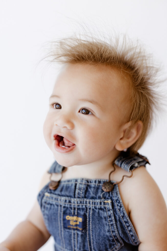 Smiling baby boy with spiky hair and expressive eyes, wearing denim overalls, captured mid-laugh during a personality portrait session in a bright studio in Roseville, CA.