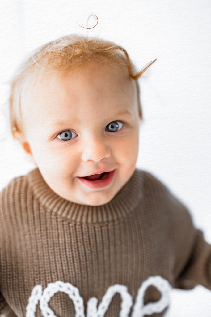 Smiling baby with blue eyes in a brown sweater, standing against a light background—perfect for milestone photography by your Sacramento family photographer.