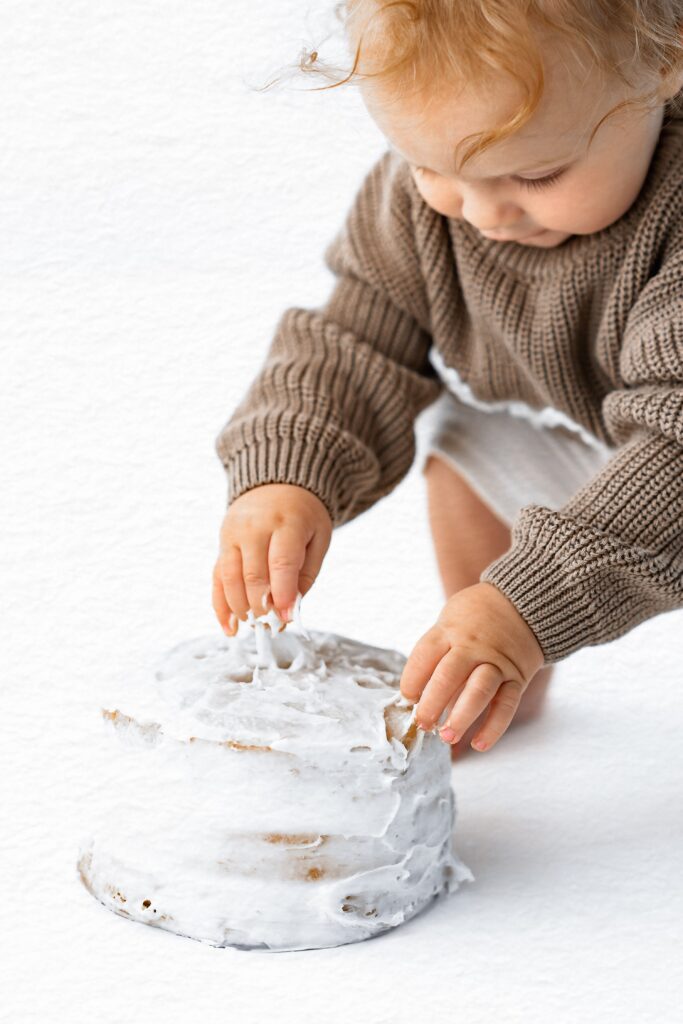 A baby in a brown sweater touches a white-frosted cake on a white surface, capturing the sweetness of milestone photography by a Roseville CA photographer.
