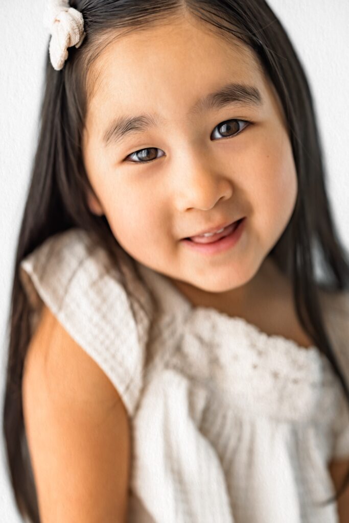 Smiling young girl with long dark hair, wearing a white dress and hair clip, captured in an in-home studio photography session against a light background.