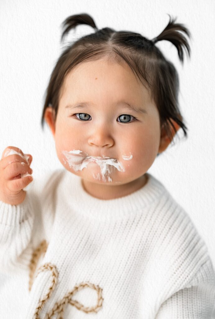 Toddler with pigtails and cream on mouth wears a white sweater, looking at the camera with a raised hand—perfect for milestone photography or babyhood sessions by a Sacramento family photographer.
