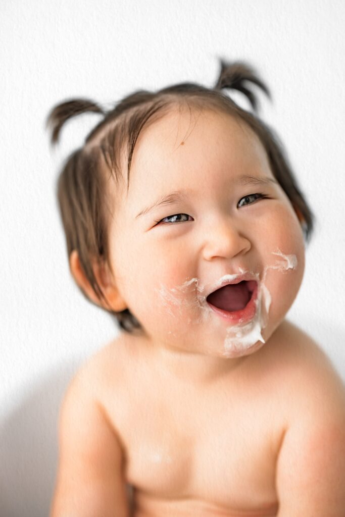 Smiling baby with pigtails and food smeared around mouth, captured in an in-home studio photography session against a plain white background.