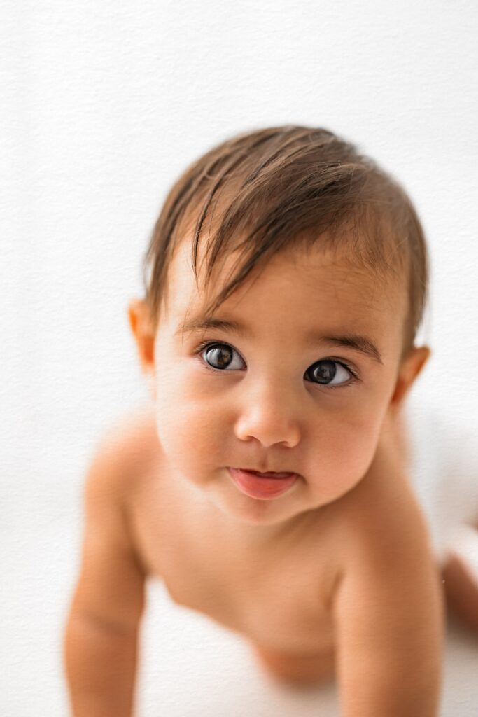 A baby with brown hair and big eyes looks up while crawling on a white background—perfect for Babyhood sessions by a Roseville CA photographer.