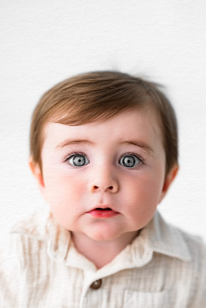 A baby with light skin, blue eyes, and brown hair, wearing a cream-colored shirt, looks at the camera during an in-home studio photography session with a Sacramento family photographer.
