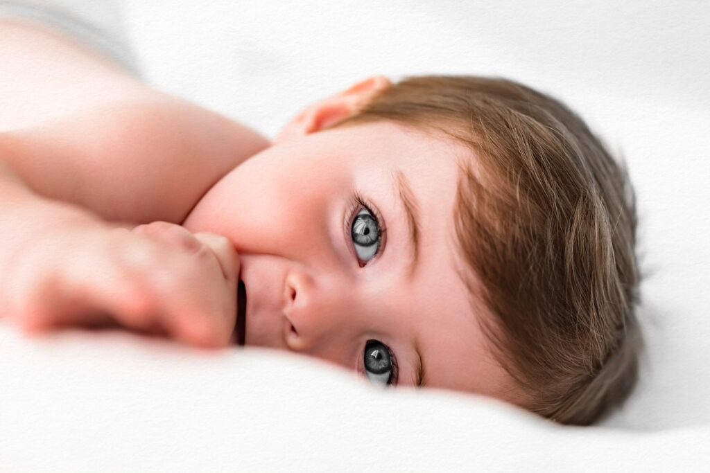 Baby with blue eyes lying on a white blanket, looking at the camera and sucking their thumb—captured during our cozy in-home studio photography Babyhood sessions.