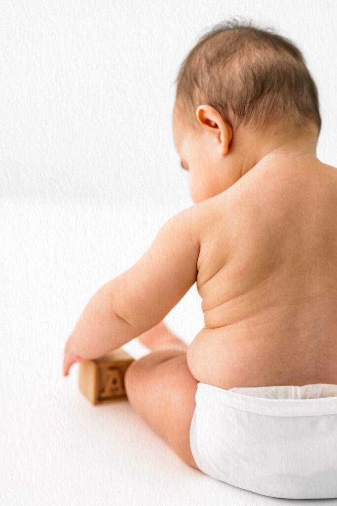 A baby in a diaper sits on a white surface, playing with a wooden block and facing away from the camera—perfect for milestone photography or an in-home studio photography session.