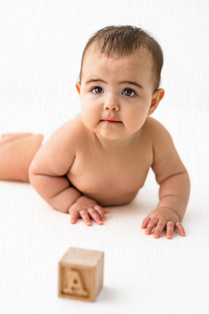 A baby lies on their stomach, looking up, with a wooden letter block in the foreground—a sweet capture perfect for milestone photography.
